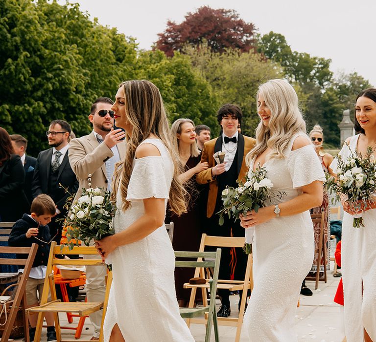 Bridesmaids walk in sparkly white dresses with front slit and white and green classic bouquets 