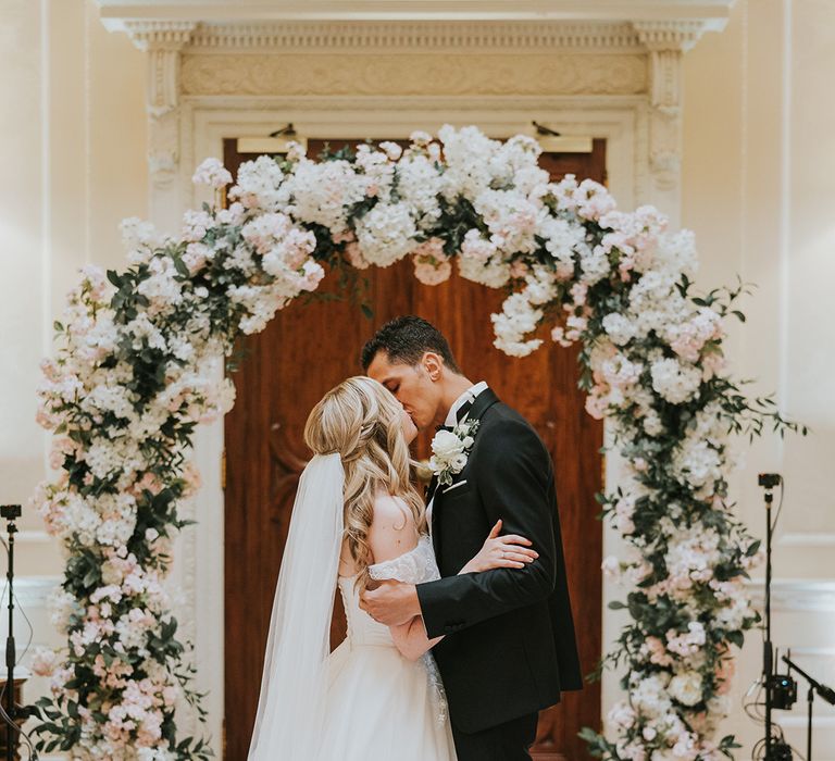 Bride and groom share their first kiss as a married couple in front of pink and white flower arch 