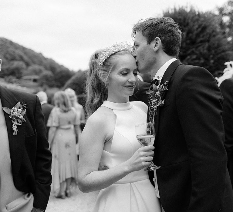 Groom in morning suit kisses the bride on the forehead as she sips from a glass 