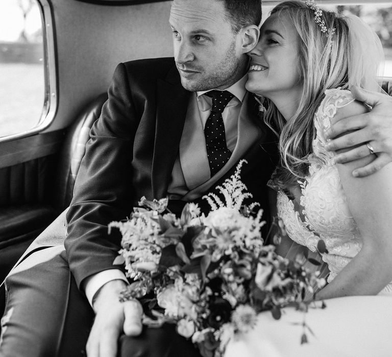 Bride in lace wedding dress wearing pearl and gold headpiece with groom in morning suit seated in vintage wedding car 