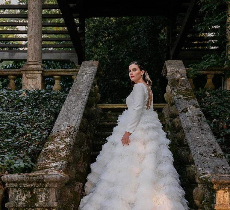 Bride stands on staircase outdoors as her long tulle skirt flows behind her 