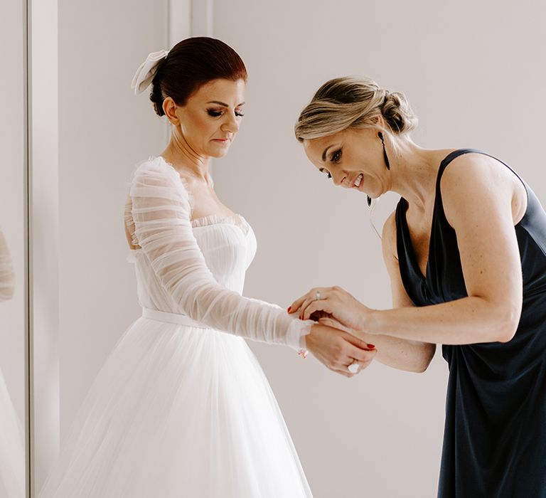 Bride in Emma Beaumont dress with ruffle detail gets help with the finishing touches from a bridesmaid in a navy blue dress