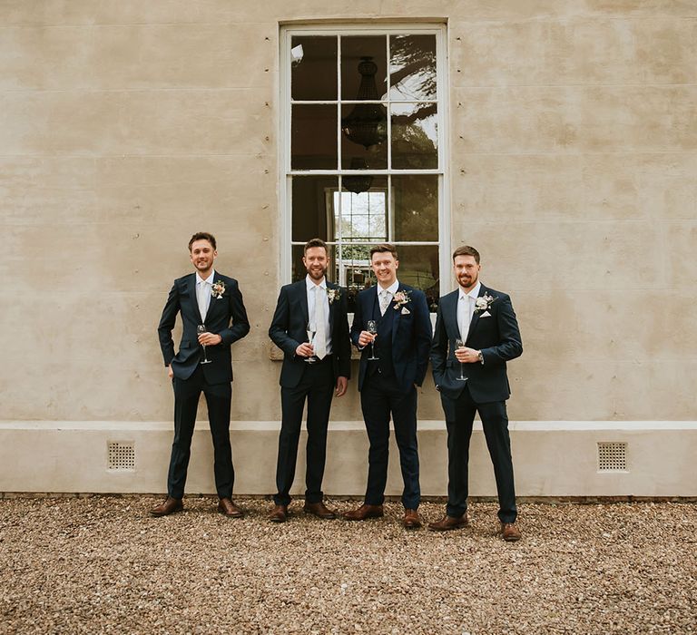 Groom stands with his groomsmen outdoors who wear blue suits with white shirts and floral buttonholes 