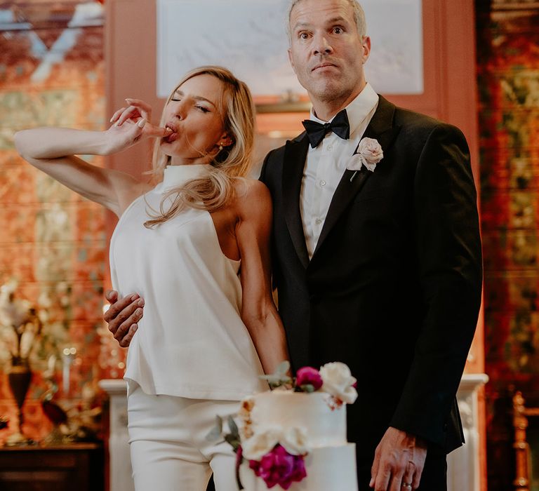 Bride licks icing off her finger from their toe tier white wedding cake decorated with pink and white flowers with the groom 