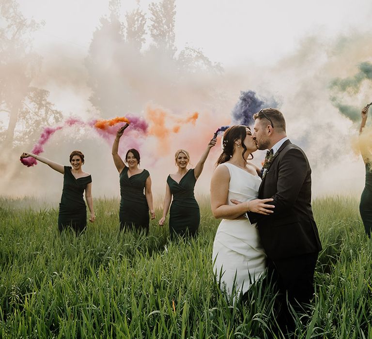 Bride and groom kiss as the wedding party wave colourful and bright smoke bombs 