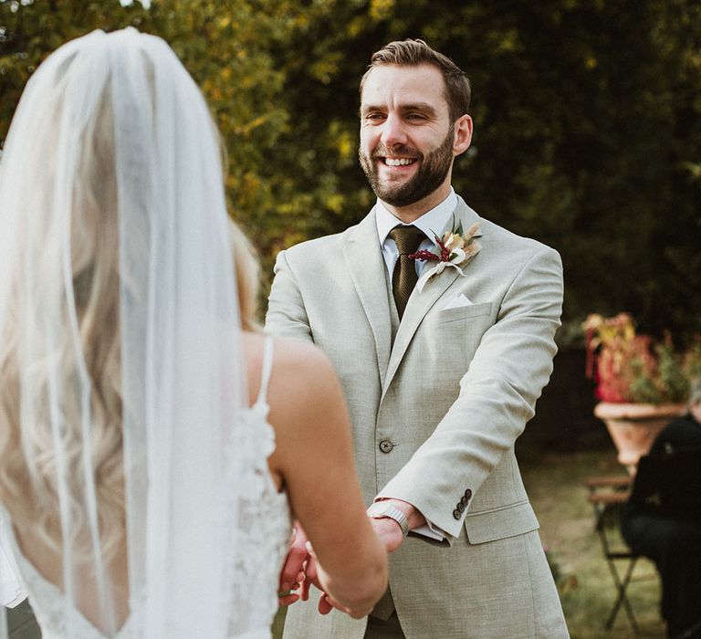 Groom wears classic stone beige linen suit, white shirt and wool green tie alongside a floral buttonhole