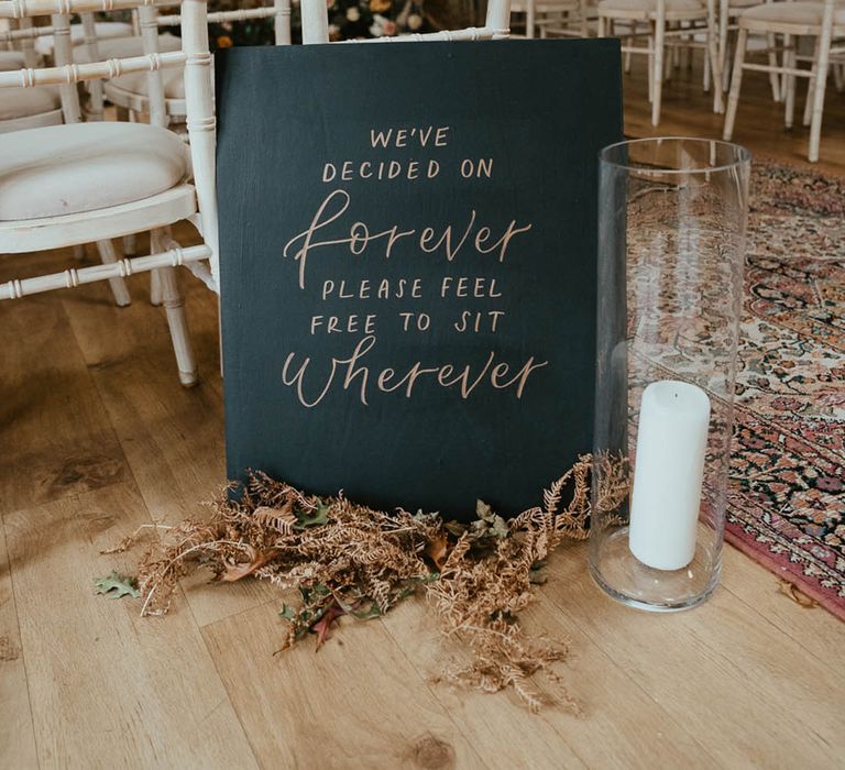 Black and gold sign with dried foliage and a white pillar candle in a hurricane vase 