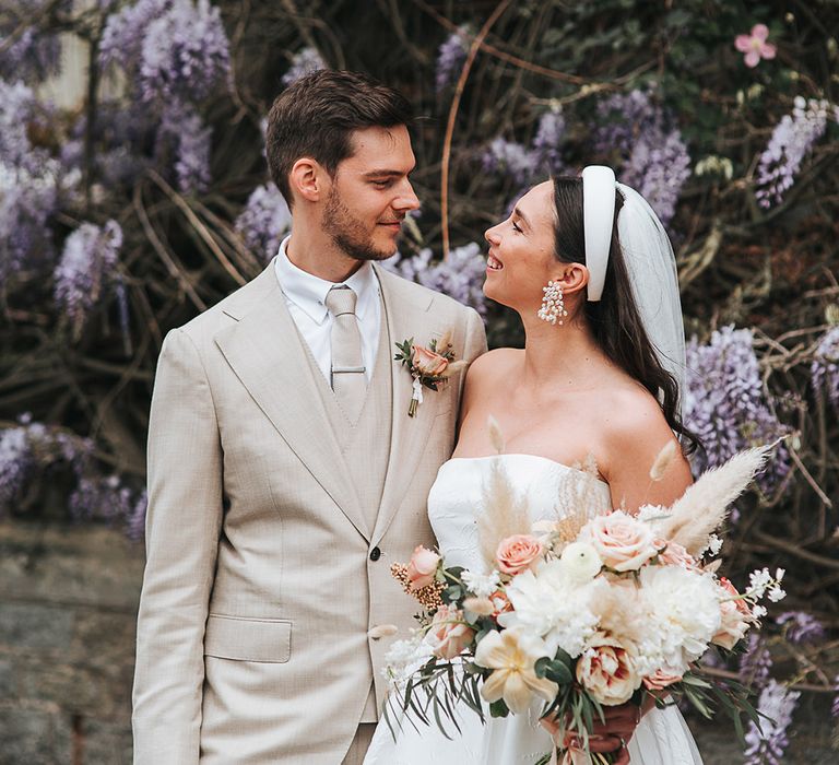 Bride with pretty accessories holding a fresh and dried grass wedding bouquet smiles up at the groom in a neutral wedding suit 