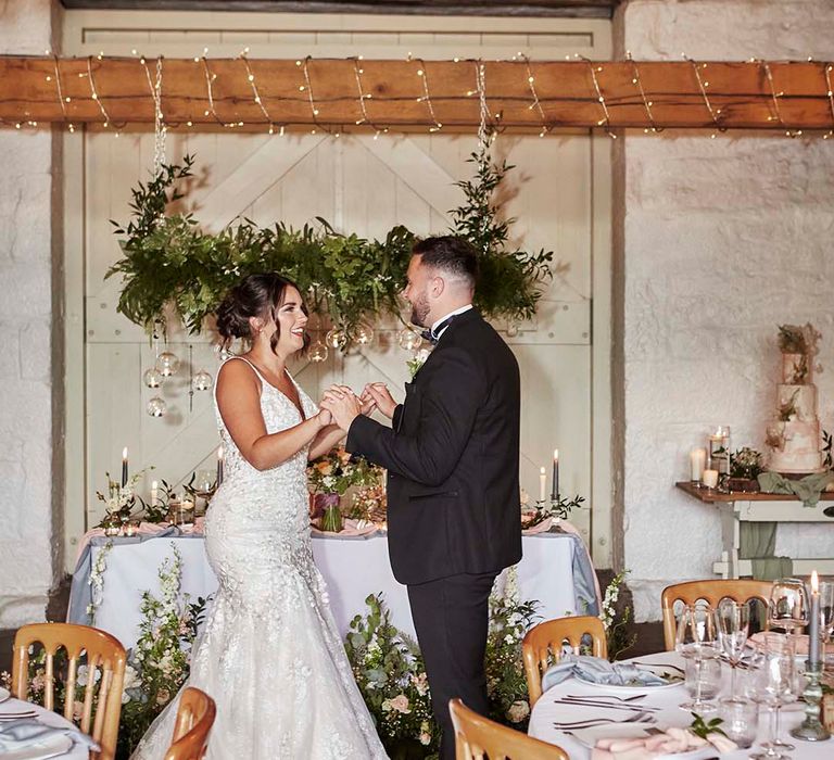 Bride in a lace fishtail wedding dress dancing with her groom in a tuxedo at their rustic barn reception in front of their sweetheart table 