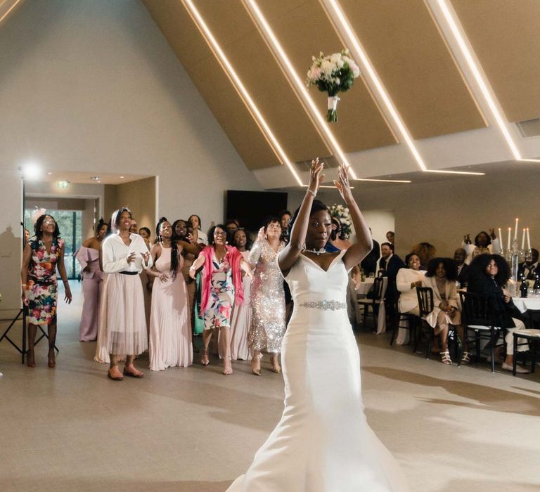 Bride wears fitted wedding dress with diamanté belt to front and throws bridal bouquet behind her on the dance floor to wedding party 