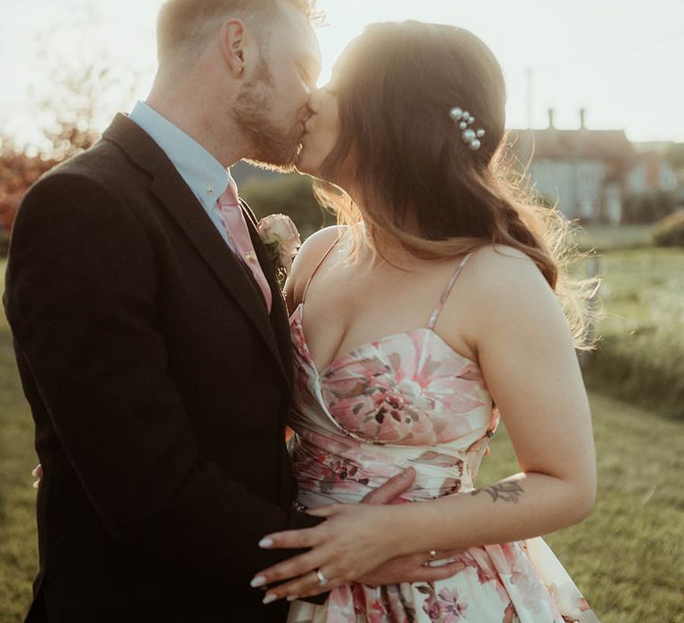 Bride with pearl hair pins with a pink flower wedding dress and groom in a brown suit share a kiss during golden hour 