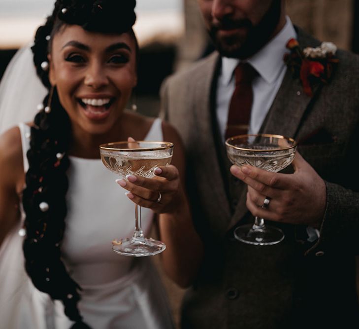 Bride holds champagne glass and laughs whilst wearing her hair in intricately braided styling complete with pearl accessories 