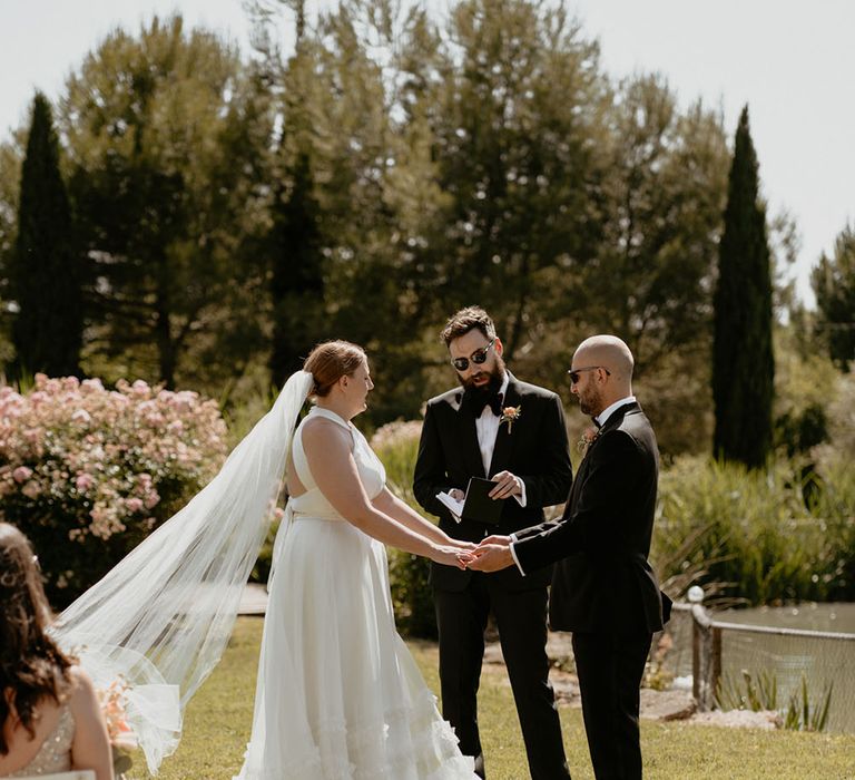 Bride's veil blows in the wind as she holds hand with her groom in black tie during outdoor ceremony in France 