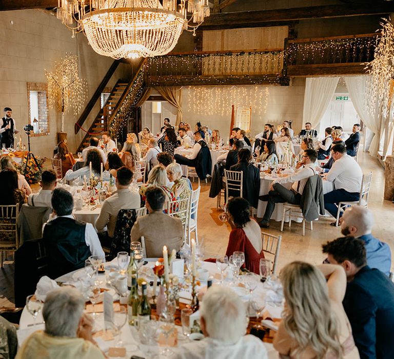 Wedding guests sit and listen to the groom's speech with a chandelier and fairy lights