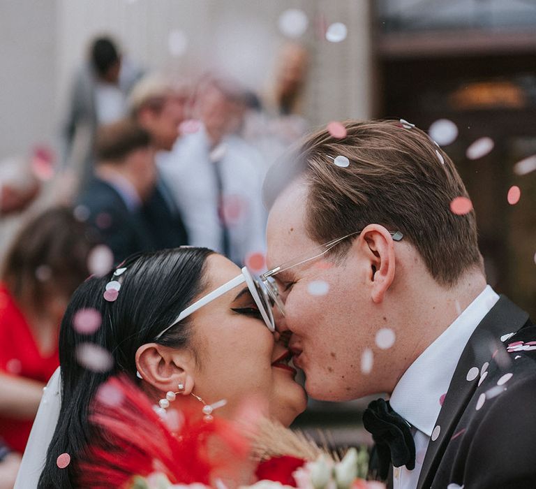 Bride wears heart-shaped sunglasses and carries bright floral bouquet as she kisses her groom whilst colourful confetti falls around them