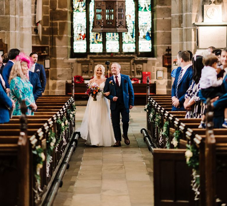 Bride walks down the aisle with her father during church ceremony whilst holding bright sunflower bridal bouquet 