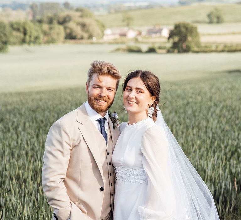 Groom in cream three piece with a dark blue tie standing in a field with the bride in a vintage wedding dress with a high neck and balloon sleeves