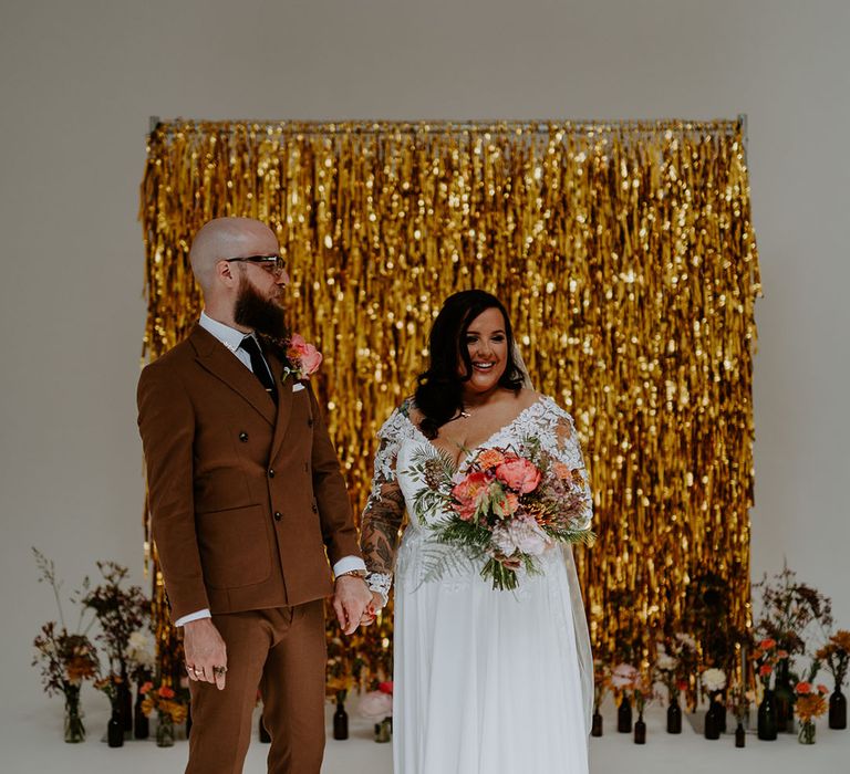 Groom wears 70s style retro brown suit and stands beside his bride in lace wedding dress holding brightly coloured floral bouquet 