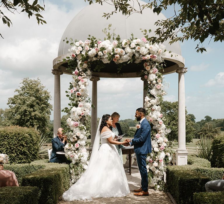 Bride & groom during outdoor Sri Lankan wedding stand beneath wedding dome finished with floral archway 