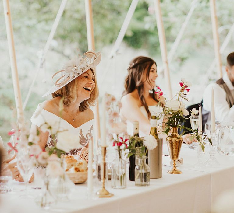 Mother of the bride in a pale pink hat and off the shoulder white wedding dress laughs at the reception 