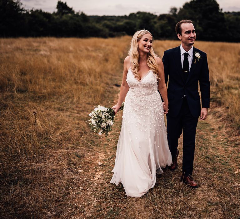 Bride in a flower appliqué Alexandra Grecco Lana wedding dress walking in the fields with the groom in a navy blue suit 