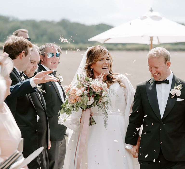 Bride in romantic wedding dress walks through white petal confetti with her groom in black tie