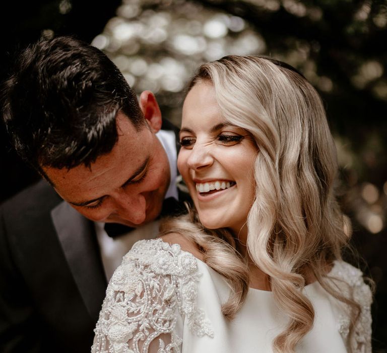 Smiling bride with curled blonde hair and beaded sleeve wedding dresses with the groom in black tie 