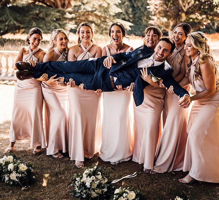 The bridesmaids in matching pink satin halter neck dresses lift the groom in a navy blue suit