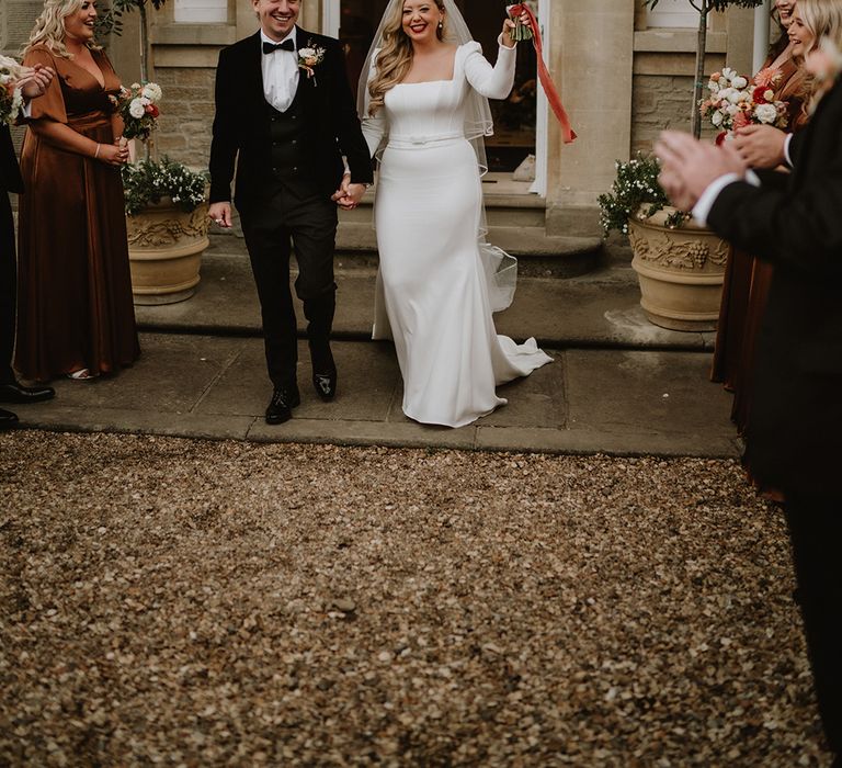 Bride in square neck long sleeve satin wedding dress with two tier veil holding colourful mixed floral bouquet with groom in black suit with floral boutonniere walking out of venue