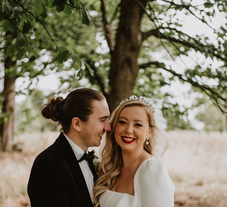 Bride in square neck corset look satin wedding dress with two tier veil laughing with groom in black suit with floral boutonniere at St Tewdrick's