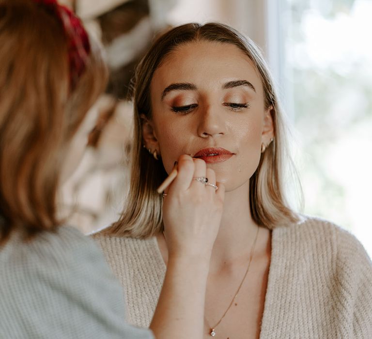 Bride gets her makeup done for her outdoor autumnal wedding with warm toned lipstick 