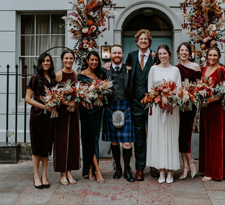 Bride in long sleeve lace vintage wedding dress, groom in dark suit with orange tie and autumnal boutonniere, best man in dark blue kilt and bridesmaids in mismatched velvet bridesmaid dresses standing underneath large floral arch with roses, peonies, foliage and pampas grass