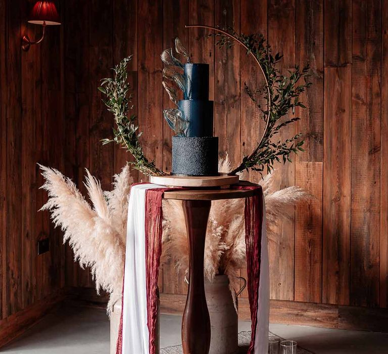 three-tier wedding cake in a rich blue buttercream with a velvet effect base tier and gold rice paper sails, finished with a semi-circle Bramble Sky arch stand and decorated with lots of dark green ruscus 