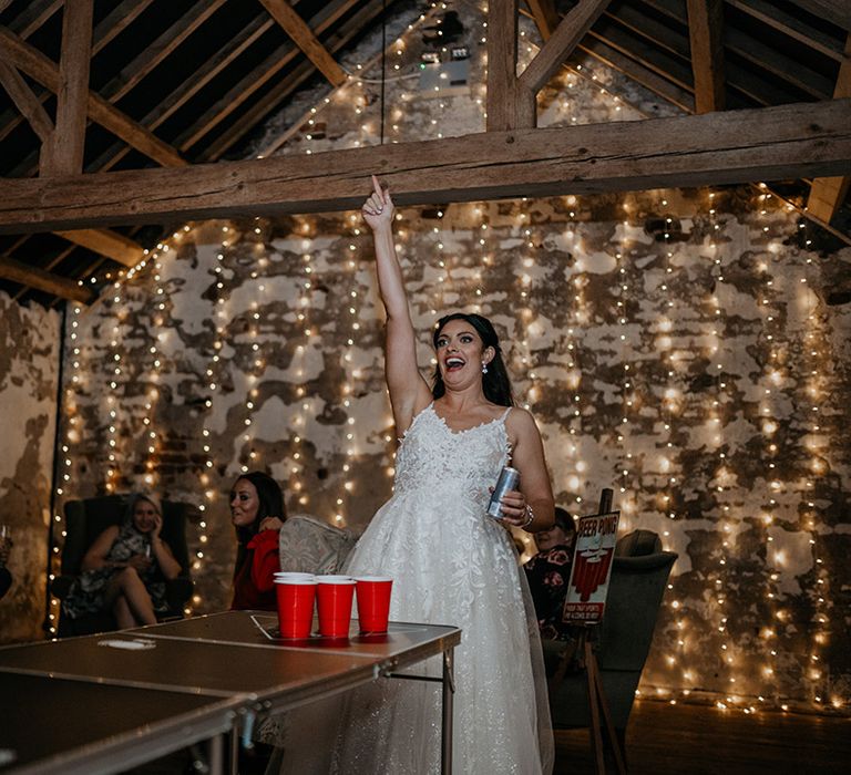 Bride in lace wedding dress celebrates as she sinks a ball for beer pong with string fairy lights for rustic barn wedding venue 