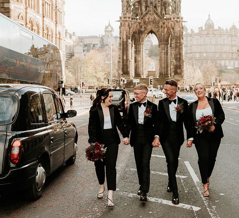 I do crew in matching outfits: two grooms in matching black velvet tuxedos with black bowties and red rose and foliage boutonnieres, the I Do Crew wearing black velvet blazers, black leather trousers and holding large red rose floral bouquets