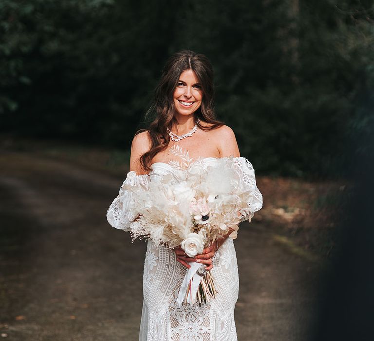 Bride in boho fitted wedding dress with a train and detachable sleeves holding an all white fired flower bouquet tied with white ribbon for boho luxe wedding 