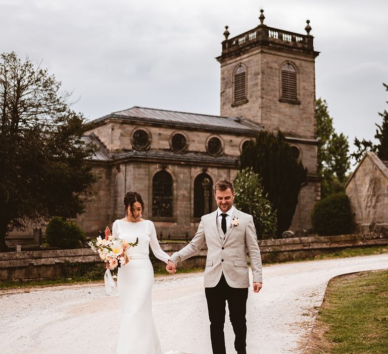 Bride in long satin wedding dress with bridal cover up semi sheer crop top holding floral wedding floral bouquet with pink garden roses, foliage and baby's-breath and groom in black suit trousers, grey blazer and light pink rose boutonniere walking around wedding venue