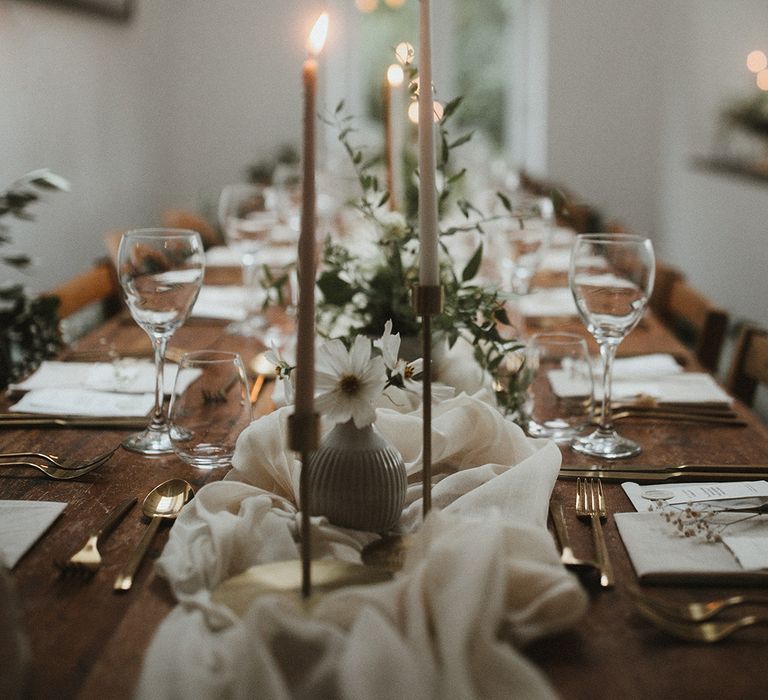 Classic tablescape across wooden tables complete with white fabric table runner and lit candles in brass holders