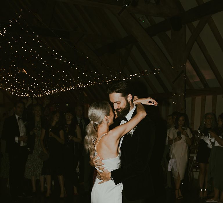 Bride with blonde hair in a sleek ponytail sharing a first dance with the groom in traditional black tie with fairy lights 