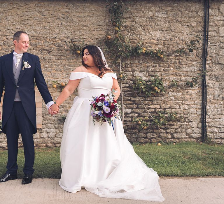 Groom in a navy blue morning suit with a white tie and white rose buttonhole with the bride in a pleated skirt dress holding a blue wedding bouquet