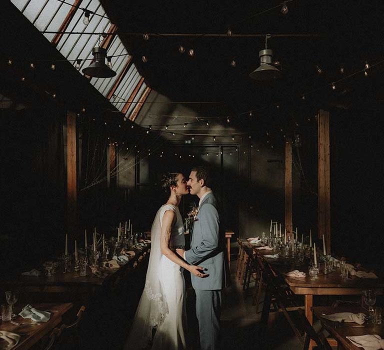 Lisa & Gabriel kiss in the middle of wooden banquet tables and industrial setting for wedding in France 