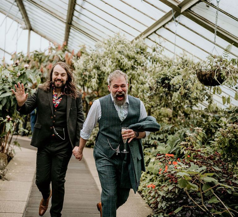 Grooms laugh and run through botanical greenhouse filled with lush greenery and florals at Kew Gardens 