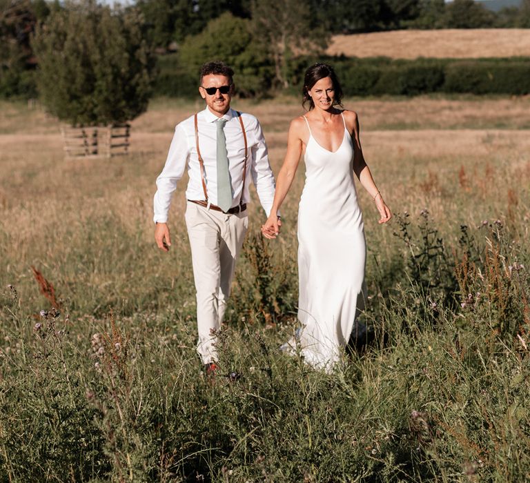 Bride in silk Grace Loves Lace wedding dress walks alongside her groom in white shirt and braces with sage green tie 