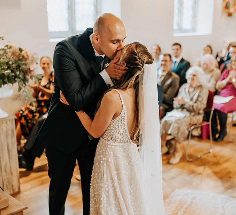 Groom in navy suit shares first kiss with bride as a married couple at their church wedding ceremony with the bride in a patterned Made With Love wedding dress