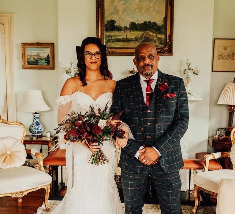 Father of the bride in three piece checkered suit with the bride in off the shoulder lace wedding dress with glasses holding dark red bouquet 