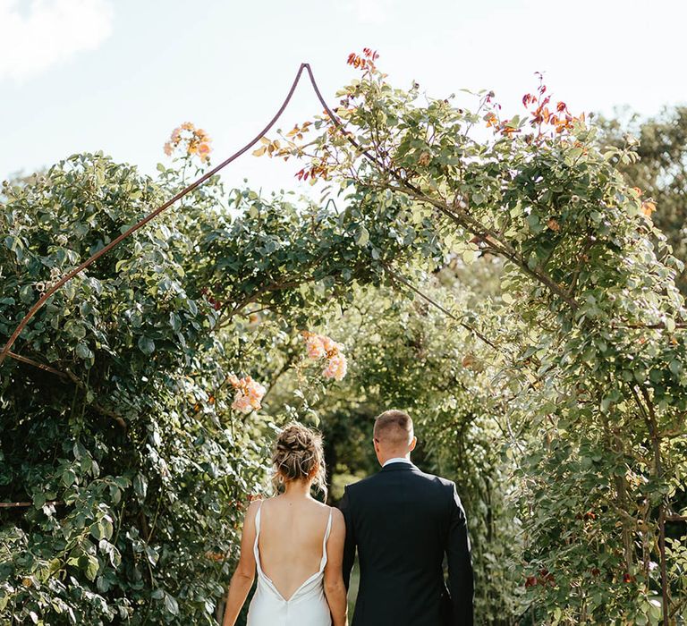 Bride in white satin wedding dress with low back detail walks with the groom outside at their rustic luxe and timeless wedding 