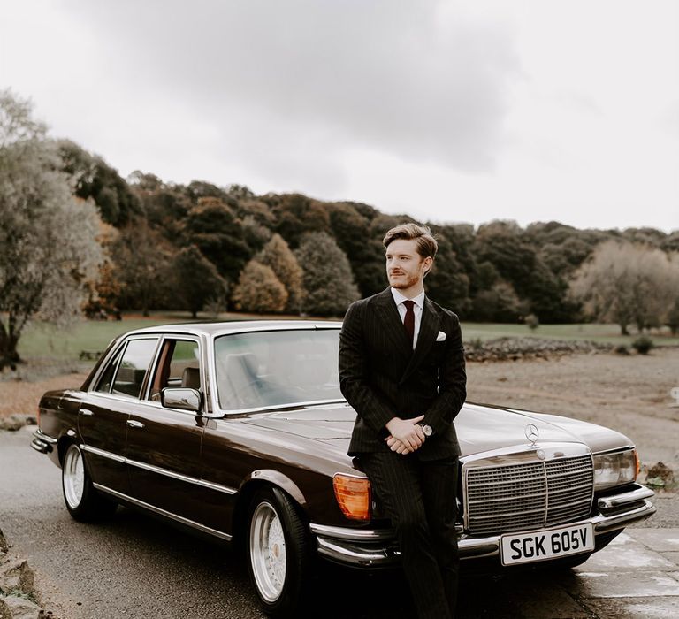 The groom in a pinstriped suit standing against the brown Mercedes wedding car 