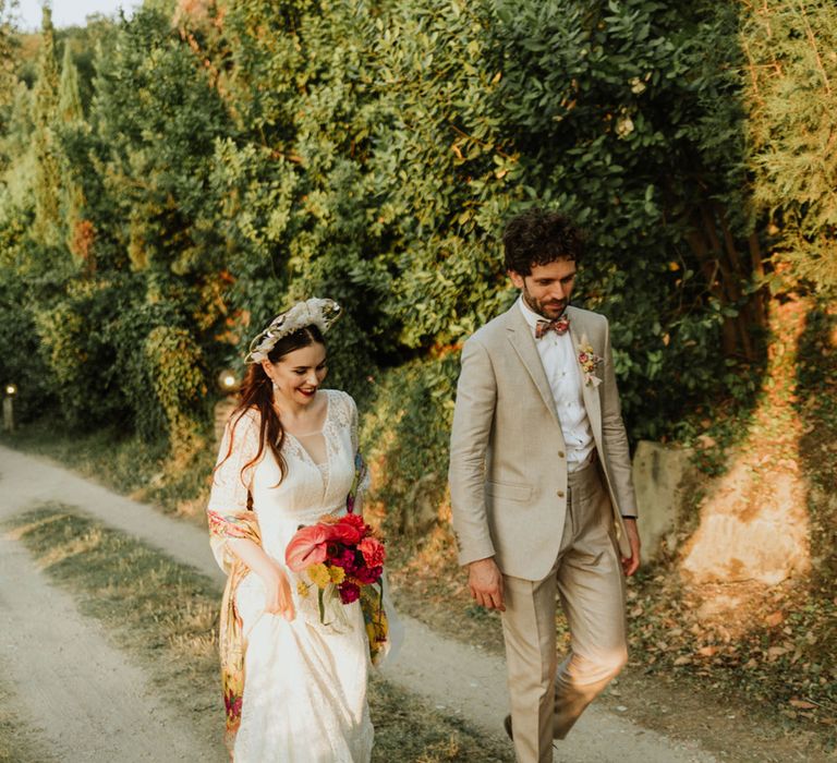 Bride and groom walk through greenery at italian destination wedding wearing white lace wedding dress, white floral structured bridal crown and greige suit