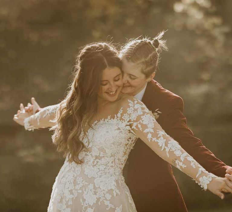 Bride in maroon bridal suit with grey waistcoat, dusky pink tie and autumnal dried flower boutonniere doing the Titanic pose with bride in long sleeve lace illusion sleeve wedding dress with overlay 