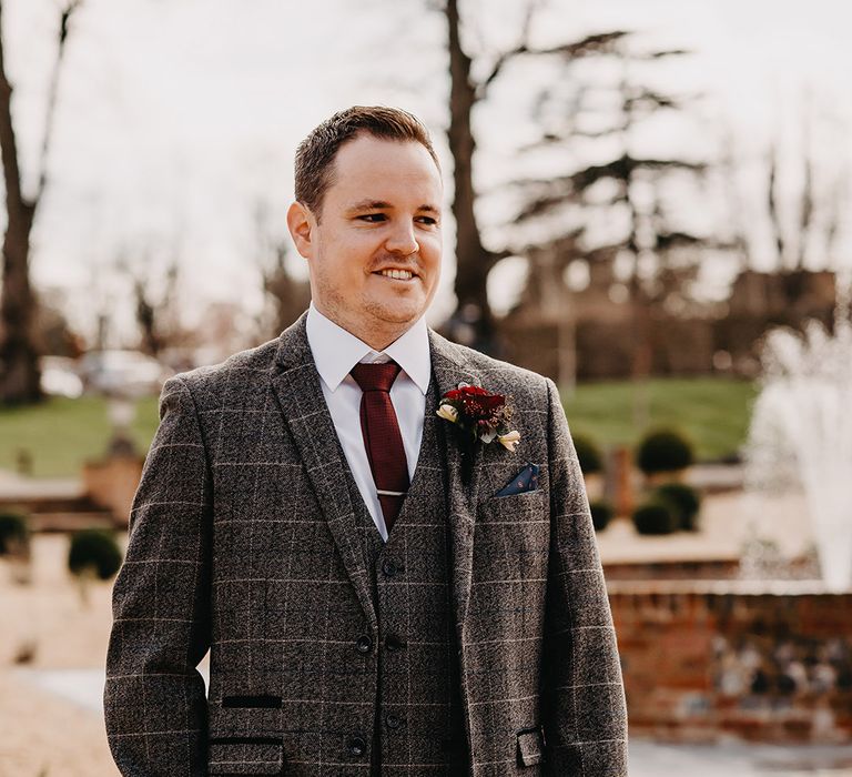 Groom in three piece grey checkered wedding suit with dark red tie and red rose buttonhole 